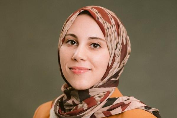 Dina Eissa in a patterned brown and beige hijab and orange top, photographed against an olive-green background.