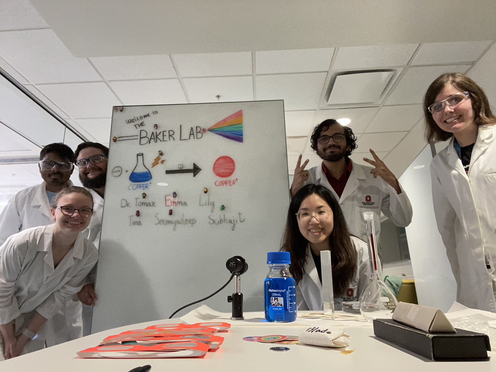 Students in lab coats standing beside a white board labeled "Welcome to the Baker Lab" with drawings of a light spectrum and a beaker
